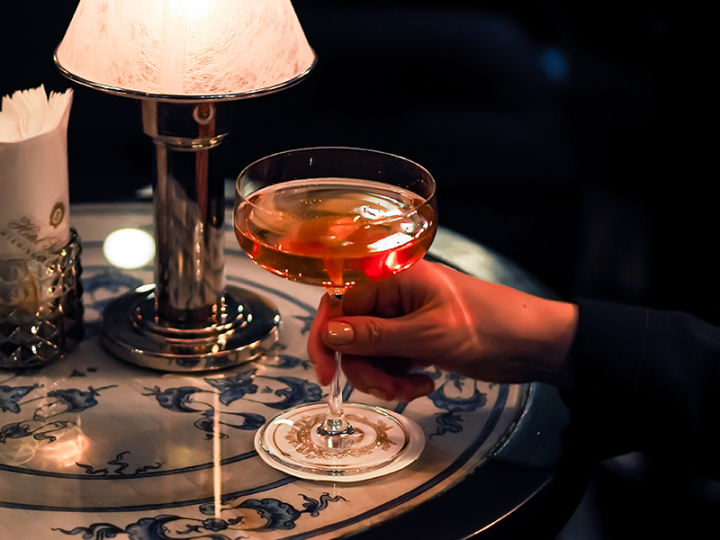 Elegantly laid table scene with a hand holding a cocktail glass, illuminated by a stylish table lamp.