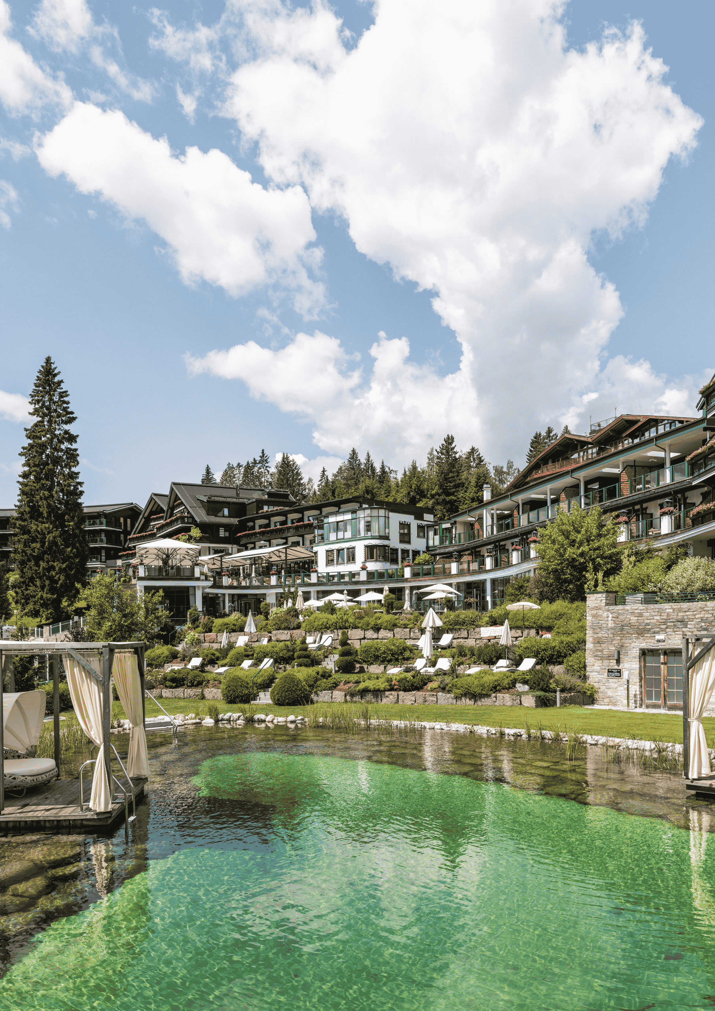Hotel Sacher Seefeld in summer: Natural swimming pond with a cabana, surrounded by lush green trees and the resort.
